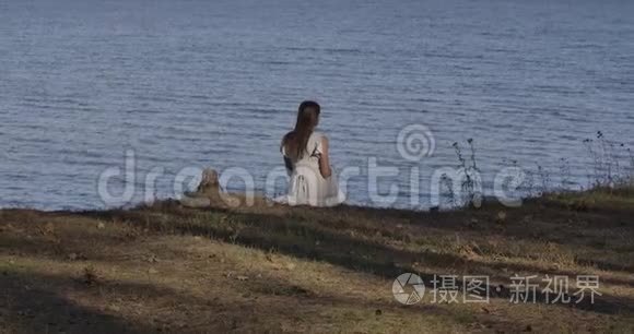 年轻悲伤的白种人女人穿着浅色的白色连衣裙坐在海滩上看着海浪。 美丽的女孩夏天