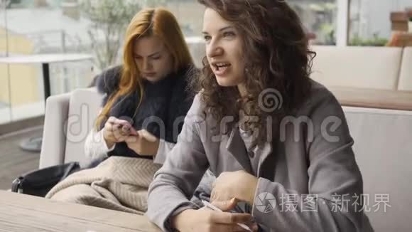 两个漂亮的白种人女朋友坐在咖啡馆的桌子旁。 一个女孩拿着电话，另两个讨论项目