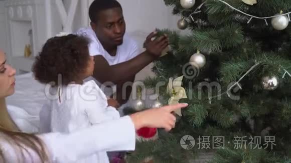 快乐的女婴在圣诞树上挂球。 孩子们装饰圣诞树。 年轻幸福的多民族家庭