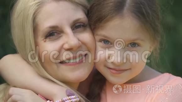 特写肖像，微笑的金发母亲和她的小女儿拥抱在夏季公园看着相机。 快乐