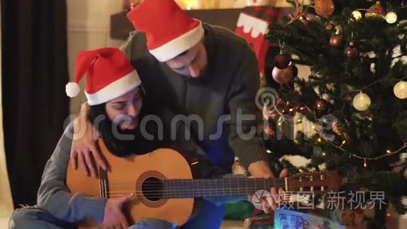 圣诞前夜圣诞老人戴`帽子的幸福夫妇的肖像。 男人教女人弹吉他。 圣诞树上的圣诞树