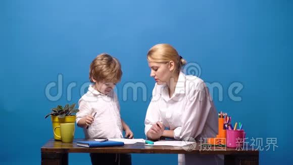 母亲坐在家里的桌子上，帮她的小儿子做蓝色背景的家庭作业。 女人和小孩