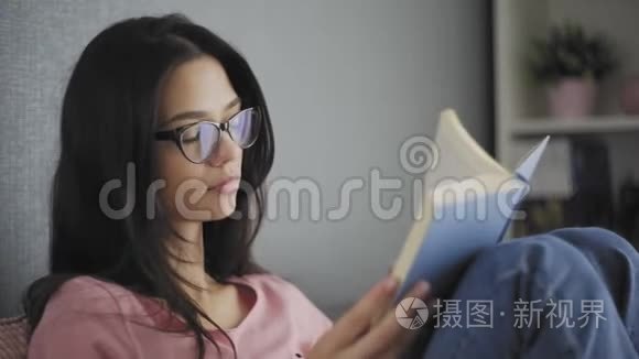 漂亮快乐的女学生戴着眼镜，在一所现代化的房子的沙发上看书。 在找。 回到过去