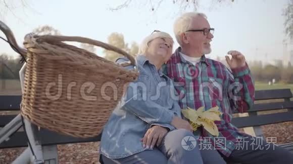侧面看白种人退休的男人和女人坐在长凳上在阳光下交谈。 成熟的欧洲家庭或