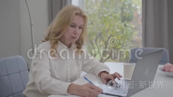 严肃成熟的白种人女性坐在家里，在网上工作。 自信的商务小姐穿着白色衬衫