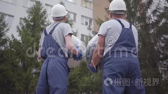 两名戴着白色头盔和蓝色制服的白人工人的背面视图。 男人互相看着