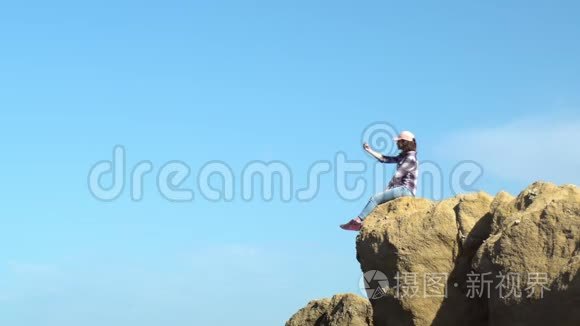 一个年轻的女人坐在一块石头上，拿着。 在岩石上自拍。 一个戴着帽子的女人在一个