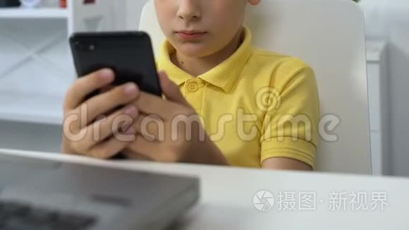 小男孩坐在笔记本电脑前，在智能手机上看视频，上瘾