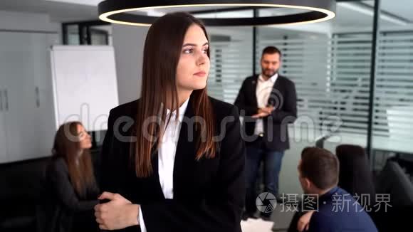 在镜头前的女商人。 她双臂交叉站在同事面前，他们坐在桌子旁