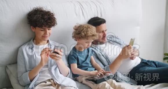 小男孩在平板电脑上玩游戏，而父母在家里的床上使用智能手机