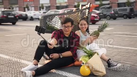 满意的年轻亚洲夫妇坐在市场手推车附近的地上，用“苹果”自拍
