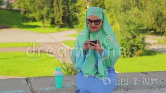 公园里一位年轻的穆斯林妇女戴着绿色围巾和太阳镜，喝着咖啡，正在送信