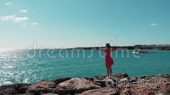 穿着红色裙子的女人，双手分开，迎着阳光站在岩石悬崖码头，享受着风和阳光。 海浪撞击岩石