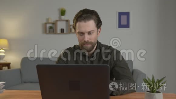 一个留着胡子的白人坐在桌子旁，在笔记本电脑上工作，在家工作，看着相机，严肃。