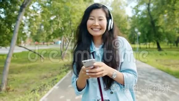 戴着耳机跳舞的亚洲女人带着智能手机在公园散步