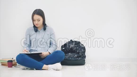 年轻的亚洲女孩坐在地板上，厌倦了写作，打哈欠。 所以，她拿着手机和耳机听着