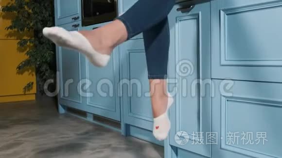 女人的腿挂着蓝色的厨房抽屉。 穿着牛仔裤和白袜子的年轻女性赤脚坐在厨房里