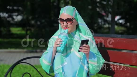 一位戴着浅色围巾和太阳镜的年轻穆斯林妇女坐在公园的长凳上，喝着咖啡，说话