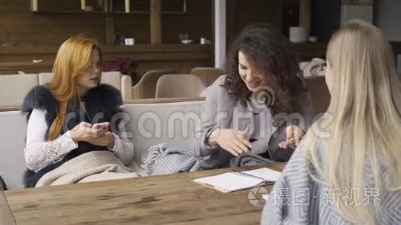 三个白种人女朋友坐在咖啡馆的桌子旁。 一个女孩拿着电话，另两个讨论项目