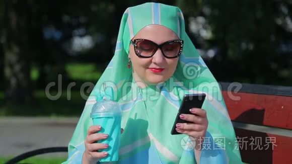 一位戴着浅色围巾和墨镜的年轻穆斯林妇女坐在公园的长凳上，喝着咖啡，在公园里聊天