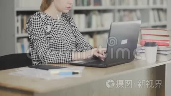 漂亮的女学生戴着耳机坐在图书馆的桌子上工作，集中在学校的项目上。