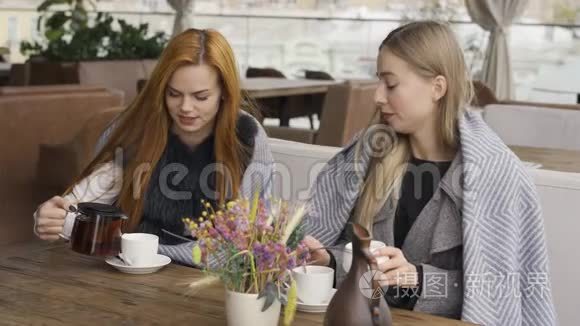 两个裹着毯子的白人女友坐在咖啡馆里聊天，喝着热茶。年轻女性放松