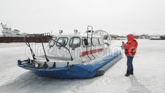 穿橙色夹克的女人在气垫船背景上使用手机。 冰冻水冬湖上的浮船。 冬季船只