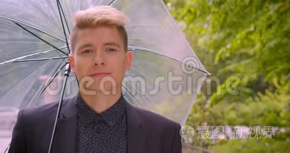 一位年轻迷人的白种人金发男子手持一把伞，笑着愉快地看着镜头的特写镜头