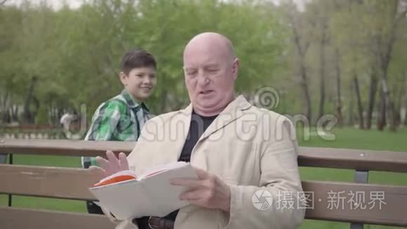 肖像可爱的秃头成熟男人坐在公园的长凳上看书。 可爱的小男孩从后面走来