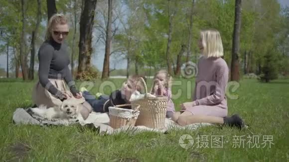 两个年轻的母亲和他们的孩子坐在绿色神奇公园的毯子上，在春天的一天。 妈妈和儿子