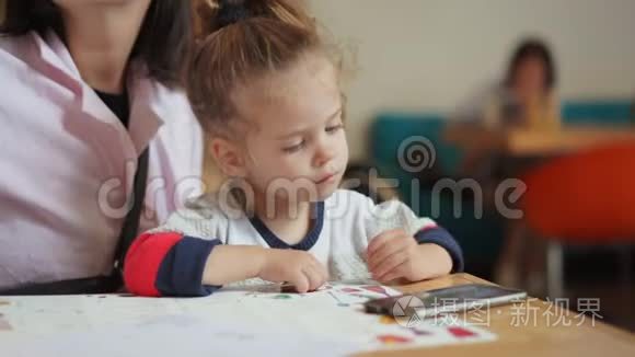 一个欧洲貌美的小女孩正坐在妈妈怀里的一张桌子上，看着智能手机上的视频