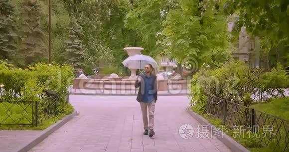 英俊的时髦商人带着雨伞微笑着走进公园里的摄像机正对着镜头。