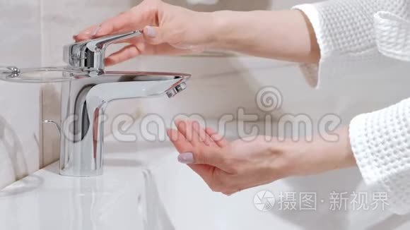 一个穿着白色长袍的女人在浴室水槽里洗手。 水从水龙头流出。