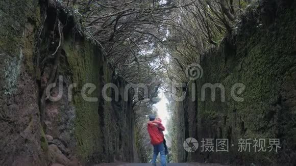 一对年轻乐观的夫妇在TenerifeAnaga自然公园的一条被树木覆盖的岩石之间的道路上。 强劲的风