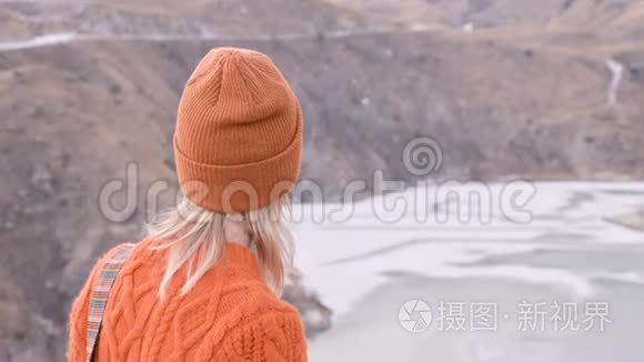 从后面看，一个穿着橙色毛衣和帽子的旅行女孩在户外看台上，背景是史诗般的背景