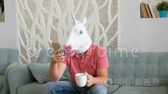 奇怪的搞笑视频：戴着独角兽面具的男人坐在沙发上拿着智能手机喝咖啡