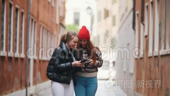 两位穿着温暖衣服的年轻女性旅行者拿着手机和相机在狭窄的街道上行走。 女人展示她的风趣