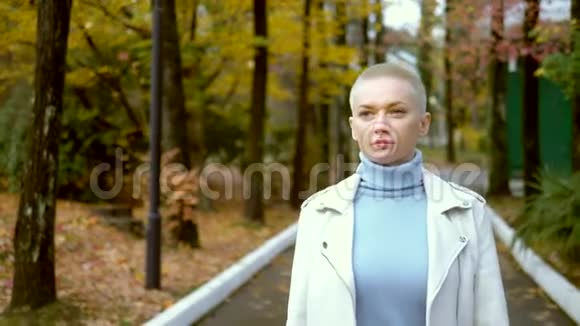 时尚的金发女人，穿着白色的皮夹克，留着很短的头发，走在秋天的公园里。 看着摄像机视频的预览图