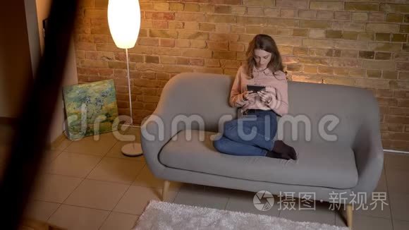 年轻的金发少女坐在沙发上，兴致勃勃地看着舒适的家中的平板电脑