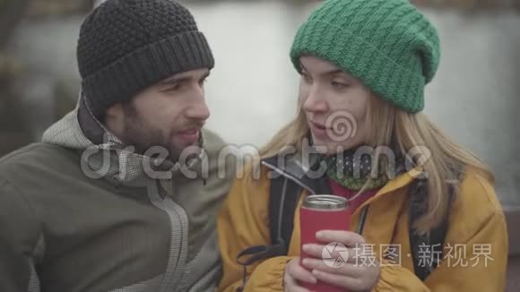 年轻夫妇穿着暖和的外套，坐在户外说话的肖像。 穿着黄色夹克和绿色帽子喝酒的女人