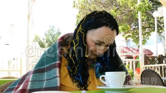 一位年长的女士，身披格子布，坐在一家夏季户外咖啡馆里。
