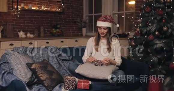 戴着圣诞帽的漂亮女孩坐在沙发上，靠近圣诞树，最后完成圣诞礼物的打包带