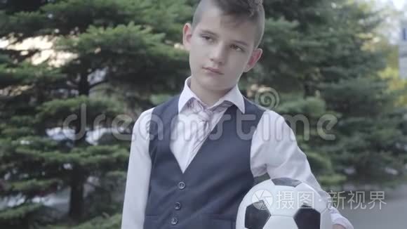 肖像可爱的衣冠楚楚的男孩站在街上，拿着足球和钱包。 严肃的年轻人