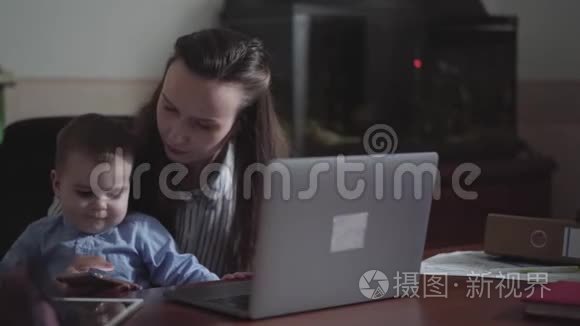 小儿子坐在桌子旁的母亲腿上。 忙碌的年轻母亲在家工作，使用手机和笔记本电脑。 孩子