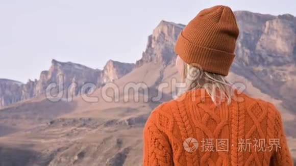 从后面看，一个穿着橙色毛衣和帽子的旅行女孩在户外看台上，背景是史诗般的背景