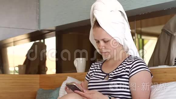一个头上戴着白色毛巾的女人拿着电话坐在床上。