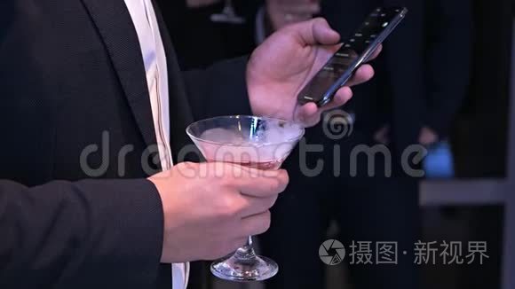 一个穿着经典西装的体面男人手里拿着一杯吸烟鸡尾酒，在聚会上用手机。 创意创意