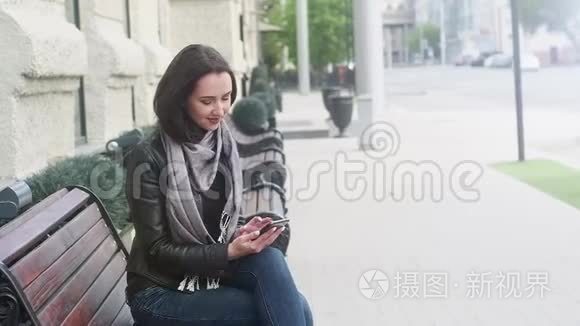 漂亮的女孩穿着黑色皮夹克和灰色围巾坐在长凳上看着电话。 他抬起头微笑着