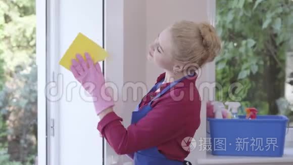 漂亮的微笑金发女孩穿着蓝色围裙，用窗户抹布在房间里洗窗户。 清洁日。 积极正面