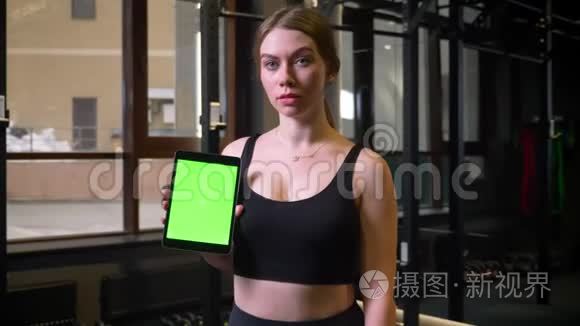女运动员展示带有彩色绿色屏幕的平板电脑进入摄像机，保持冷静，集中在健身房，